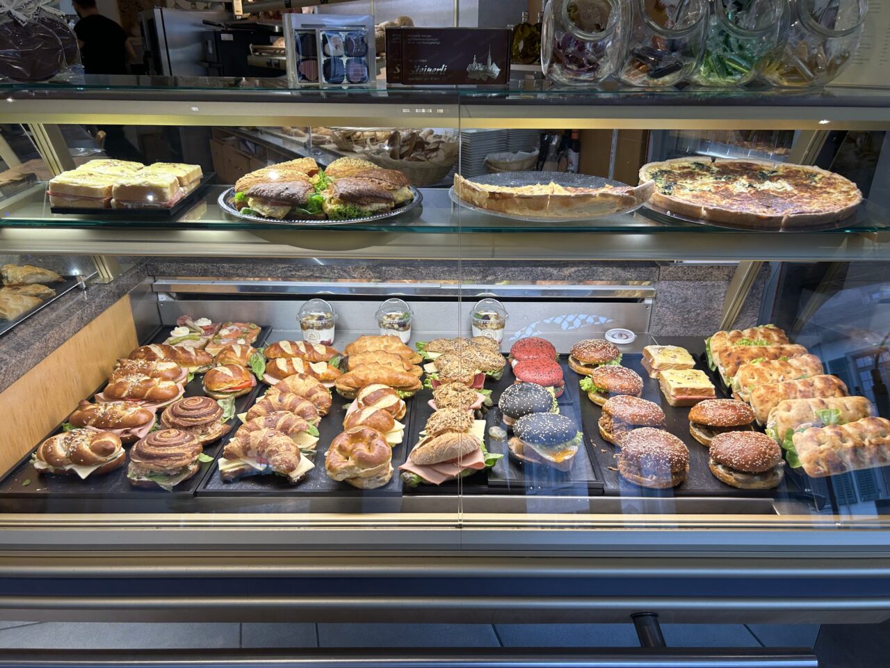 Vitrine mit vielfältigen Backwaren: Sandwiches, Brötchen, Quiche und Gebäck, frisch zubereitet in einer Bäckerei.